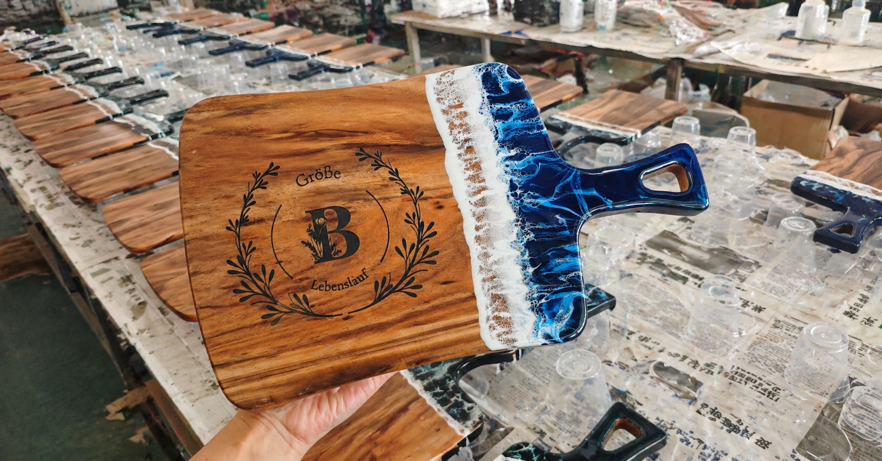 Handcrafted acacia wood serving board with blue ocean wave resin overlay, custom laser-engraved monogram, held in artisan workshop production line